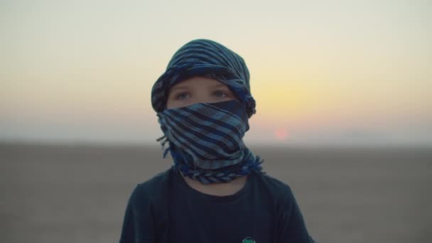 Retrato de un niño de 5 años con keffiyeh a cuadros azules parado en el desierto al amanecer. Desierto amanecer con niño turista mirando a la cámara. — Vídeos de Stock