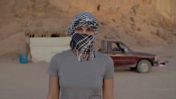 Retrato de una mujer vestida de blanco y negro a cuadros keffiyeh de pie en el desierto mirando a la cámara. Caucásico mujer turista quitándose el tocado y sonriendo a la cámara. — Vídeos de Stock