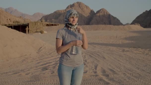 Vrouw in geruite keffiyeh drinkwater van plastic fles staat in de woestijn. Blanke vrouwelijke toerist lessen dorst tijdens woestijn avontuur. — Stockvideo