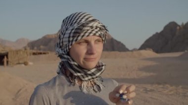 Kareli keffiyenin içinde çölde duran plastik şişeden su içen bir kadın. Beyaz kadın turist çöl macerasında susuzluğunu gideriyor. 4K