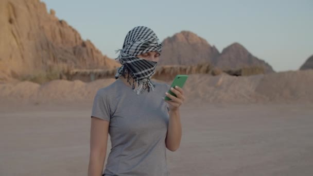 Turista femenina en keffiyeh a cuadros tratando de encontrar la red móvil utilizando el teléfono inteligente de pie en el desierto. Mujer buscando señal móvil para hacer una llamada en el desierto. — Vídeos de Stock