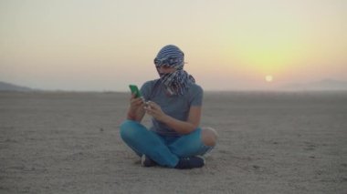 Checkered Kefiyeh 'de cep telefonuyla çölde oturan bir kadın. Çölde arama yapmak için telefon sinyali arayan bir turist.. 
