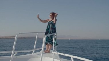 30 'lu yaşlarda bir kadın cep telefonuyla botun burnunda fotoğraf çekiyor. Elbiseli bir kadın beyaz yatta selfie çekmeyi sever..