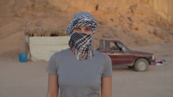 Portret van een vrouw met zwart-wit geruite keffiyeh in de woestijn kijkend naar de camera. Blanke vrouwelijke toerist opstijgen hoofdtooi en glimlachen op camera. — Stockvideo