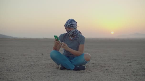 Женщина в клетчатой keffiyeh пытается найти мобильную сеть с помощью мобильного телефона, сидя в пустыне. Турист ищет мобильный сигнал, чтобы позвонить в пустыню. — стоковое видео