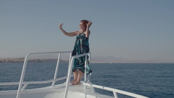 Mujer de 30 años tomando fotos en el teléfono móvil de pie en la nariz del barco. Mujer en vestido disfruta de la vela de mar haciendo selfie imágenes en yate blanco. — Vídeos de Stock