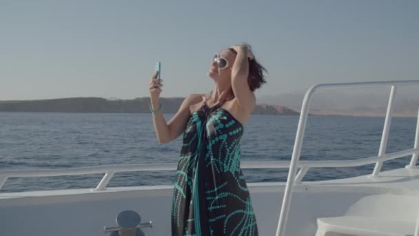 Mujer de 30 años en gafas de sol tocándose el pelo mirando a la pantalla del teléfono móvil como un espejo navegando en barco. Mujer en vestido disfrutar de la vela de mar haciendo selfie fotos. — Vídeos de Stock