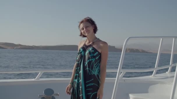 Portret 30-letniej uśmiechniętej kobiety w sukience stojącej na białym jachcie patrzącej w kamerę. Kobieta lubi rejs statkiem po morzu. — Wideo stockowe