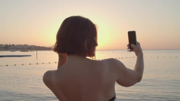 Γυναίκα που βγάζει selfie την ανατολή του ηλίου πάνω από τη θάλασσα χρησιμοποιώντας το κινητό τηλέφωνο. Πάνω από τον ώμο της πίσω όψη της γυναίκας λήψη selfie μνήμες φωτογραφία στο κινητό τηλέφωνο στέκεται στην παραλία. — Αρχείο Βίντεο