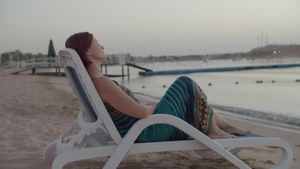 Vrouw in jurk zittend op strandstoel aan de kust kijkend naar zonsopgang boven de zee. Vrouwelijke toerist alleen op het strand. — Stockvideo