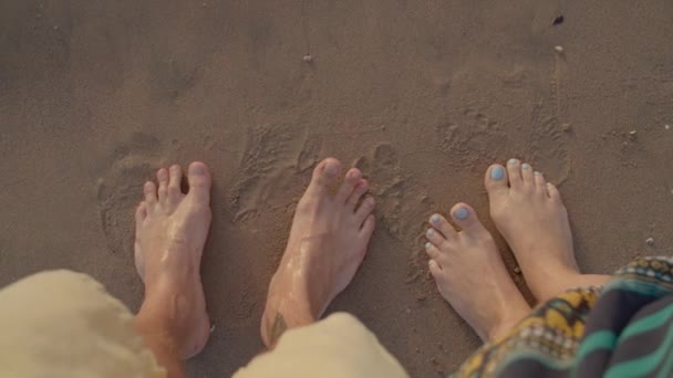 Couples feet standing on the sea shore drawing heart shape on sand. Husband and wife barefoot draw heart on the beach. Sea wave washes away painted heart on sand. — Stock Video