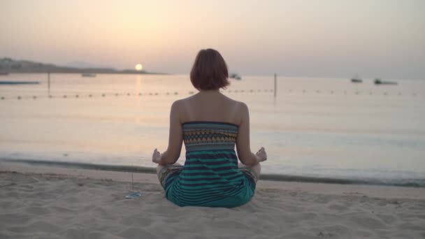 Mujer morena de 30 años en vestido sentada en posición de loto y meditando en la playa en la orilla del mar observando el amanecer sobre el mar. Vista posterior de la hembra hace práctica de respiración por mar. Concepto de salud mental. — Vídeo de stock