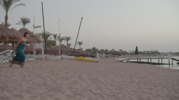 Vrouw in jurk trekt strandstoel naar de zee. Vrouw met chaise lounge om het naast de zee te zetten. dame met strand bed alleen op het strand vroeg in de ochtend. — Stockvideo