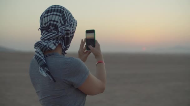 Mujer en keffiyeh a cuadros tomando fotos de la salida del sol en el teléfono móvil de pie en el desierto. Turista mujer caucásica disfrutar de la aventura del desierto. — Vídeos de Stock