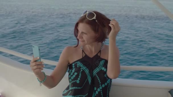 Kobieta po trzydziestce dotykająca włosów patrząca na ekran telefonu komórkowego jak na lustro pływające na łodzi. Kobieta w sukience cieszyć morze żeglarstwo co selfie zdjęcia. — Wideo stockowe