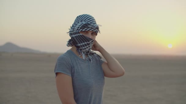 Een vrouw van in de 30 met zwart-wit geruite keffiyeh die in de woestijn staat en naar de camera kijkt. Blanke vrouwelijke toerist die haar hoofdtooi afdoet en glimlacht naar de camera. Woestijnzonsopgang. — Stockvideo
