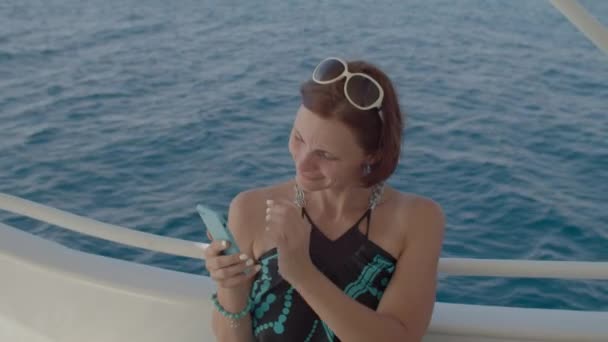 Een vrouw met een smartphone die op een boot vaart. Vrouw in jurk genieten van zee zeilen het maken van foto 's. Glimlachende vrouw in cel. — Stockvideo