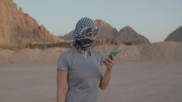 Turista trentenne in keffiyeh a scacchi che parla sul cellulare in piedi nel deserto. Donna che usa la cella nel deserto e beduino con cammello che cammina. — Video Stock