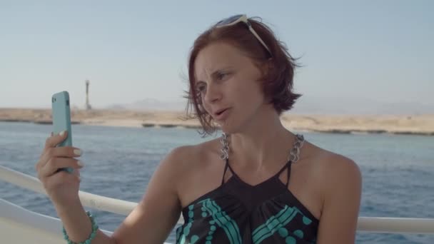 Mujer vestida hablando en video conferencia navegando en barco de mar usando teléfono celular. Mujer charlando en la videollamada sosteniendo el teléfono móvil en las manos sentado en el yate. — Vídeos de Stock