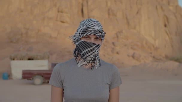 Een vrouw van in de 30 met zwart-wit geruite keffiyeh die door de woestijn loopt en naar de camera kijkt. Blanke vrouwelijke toerist opstijgen hoofdtooi en glimlachen op camera. — Stockvideo
