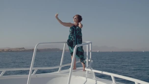 Kobieta robiąca zdjęcia komórce stojącej na nosie łodzi. Kobieta w sukience cieszy żeglarstwo morskie co selfie zdjęcia żeglarstwo na białym jachcie. — Wideo stockowe