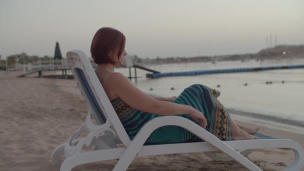 Mujer vestida sentada en una silla de playa en la orilla del mar observando el amanecer sobre el mar. Turista sola en la playa. — Vídeo de stock