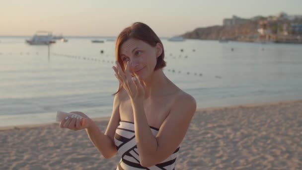 站在海滩上的女人在脸上涂防晒霜.女人用防晒霜润肤.清早一个人在海滩上. — 图库视频影像