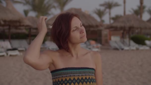 Een vrouw uit de 30 die op een leeg strand naar de zonsopgang kijkt. Bruin haar vrouw in jurk op zee kust met palmbomen. — Stockvideo
