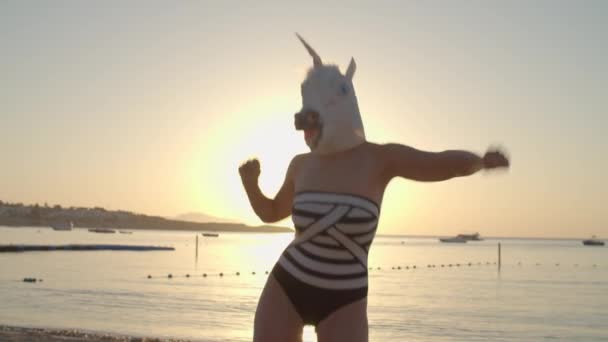 Positivo ballare in maschera testa di unicorno sulla spiaggia all'alba. Divertente danza di donna che indossa costume da bagno e maschera di unicorno. Buone vacanze. — Video Stock