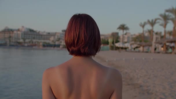 Mujer de 30 años en traje de baño caminando en la orilla del mar y viendo el hermoso amanecer sobre el mar. La hembra disfruta del amanecer en la playa. Vista posterior de la mujer que camina. — Vídeo de stock