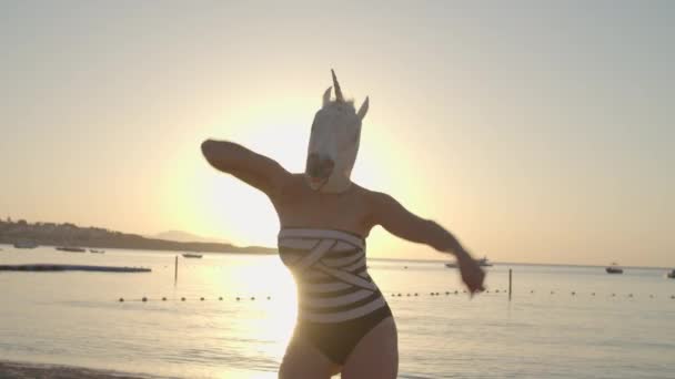 Mulher de fato de banho e máscara de unicórnio dança na praia ensolarada. Dança engraçada de pessoa usando máscara de unicórnio. Pessoas felizes. — Vídeo de Stock