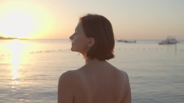 Mulher de 30 anos de maiô caminhando até o mar, voltando-se para a câmera e sorrindo. A fêmea gosta de amanhecer na praia. Visão traseira da mulher indo nadar ao nascer do sol. — Vídeo de Stock