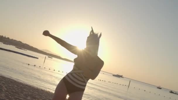 Lustige Frau mit Einhorn-Kopfmaske, die im Morgengrauen am Strand tanzt. Fröhlicher Tanz der Frau in Badeanzug und Einhornkopf. Frohe Feiertage. Kamerafahrten. — Stockvideo
