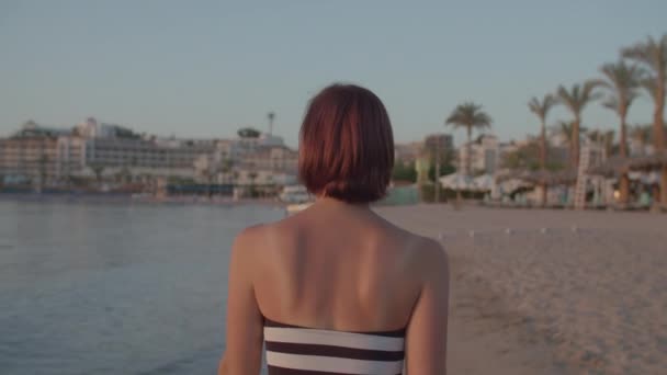 Kobieta po trzydziestce w stroju kąpielowym spacerująca po brzegu morza, zwracająca się do kamery i uśmiechnięta. Kobieta cieszy się świtem na plaży. Widok z tyłu kobiety spacerującej. — Wideo stockowe