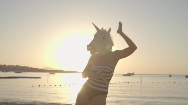 Positivo ballare in maschera testa di unicorno sulla spiaggia all'alba. Divertente danza di donna che indossa costume da bagno e maschera di unicorno. Buone vacanze. — Video Stock