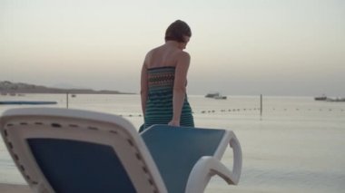 Plaj sandalyesini denize çeken elbiseli bir kadın. Dişi, şezlongla denize gidip oturuyor. Sabahın köründe sahilde tek başına dinlenen bir kadın..