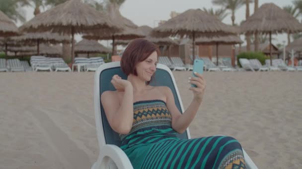 웃는 여자, 해변 의자에 앉아 핸드폰을 사용하는 여자. 빈 해변에서 핸드폰을 사용하는 여성. — 비디오