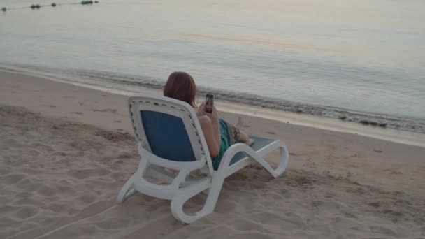 Frau macht Selfie-Foto am Meer sitzend mit Handy. Über die Schulter Rückenansicht einer Frau, die Selfie-Foto-Erinnerungen auf dem Handy macht, sitzend auf einem Strandkorb. — Stockvideo