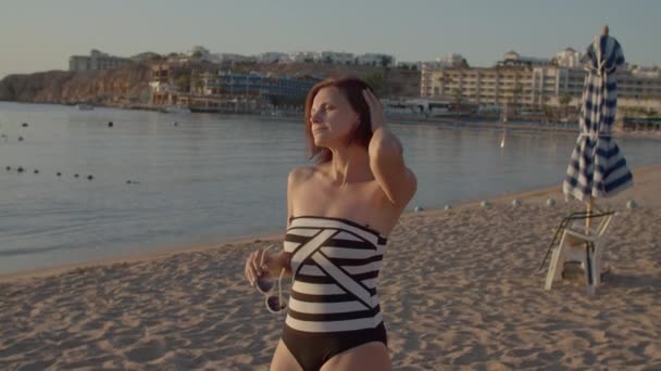 Mujer de 30 años en traje de baño de pie en la playa y con gafas de sol mirando al amanecer sobre el mar. Temprano en la orilla del mar. Persona sola en la playa. — Vídeo de stock