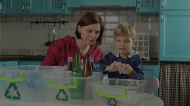 Jonge moeder en voorschoolse zoon die thuis afval sorteren. Familie sorteerpapier, glas en plastic recycleerbare materialen in containers met groene recyclingbord. — Stockvideo
