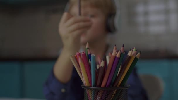 Niño en auriculares dibujo con lápices de color sentado solo en casa. Niño rubio pintando con lápices. — Vídeos de Stock