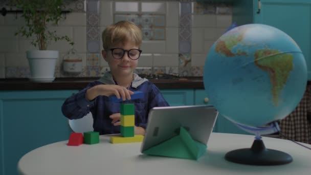 Edificio de niños de escuela con bloques de madera de color mirando a la computadora de la tableta sentada en casa. Niño inteligente haciendo asignación en línea. Niño jugando juego con constructor de madera. — Vídeos de Stock