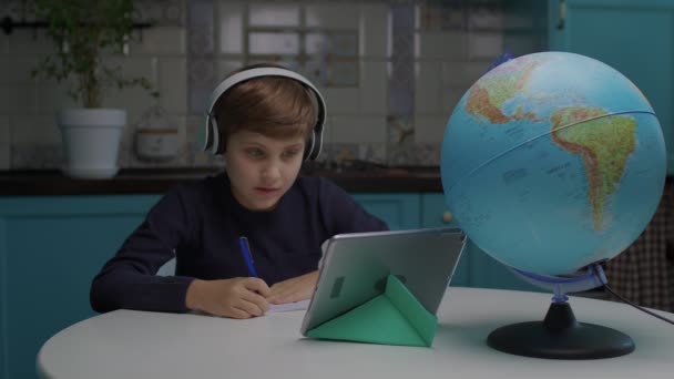 School boy with autism wearing headphones writing with pen learning online using tablet computer at home. Smiling autistic school kid studying from home. — Stock Video