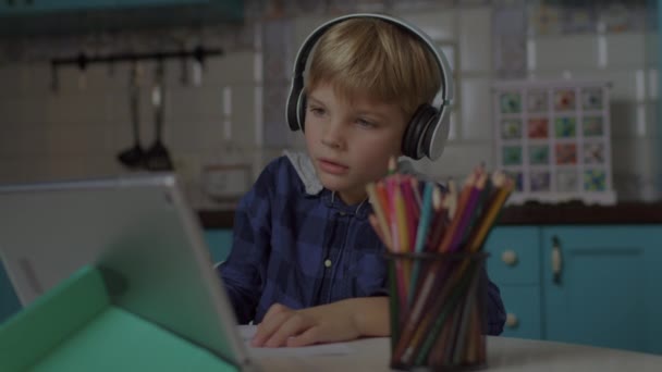 Iskolás fiú fülhallgatóban, aki online művészeti órát tart táblagéppel. Szőke fiú rajz színes ceruzák, hogy az online házi feladat. — Stock videók