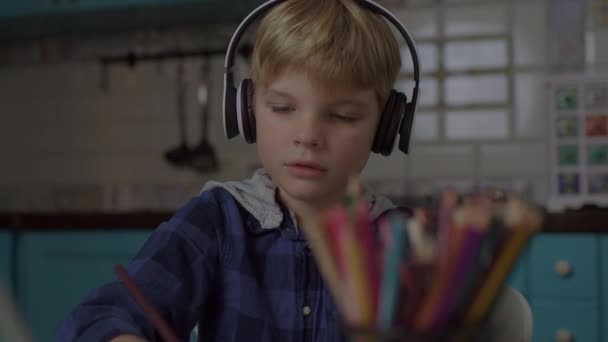 タブレットコンピュータ上のオンラインアートレッスン中にカラー鉛筆でヘッドフォンの図面で子供。ブロンドの男の子作るオンライン宿題で鉛筆. — ストック動画