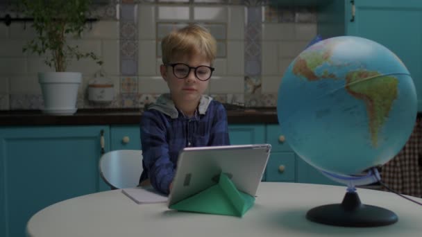 自宅からの距離学習のためにタブレットコンピュータを使用してオンラインクラスを持つ眼鏡でかわいい学童。子供がガジェットを見て話す. — ストック動画