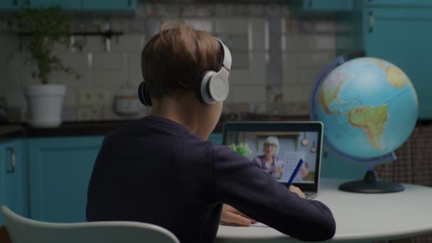 Baksida av skolan pojke i hörlurar studerar online med senior lärare på bärbar dator. Kid lärande via internet sitter hemma. — Stockvideo