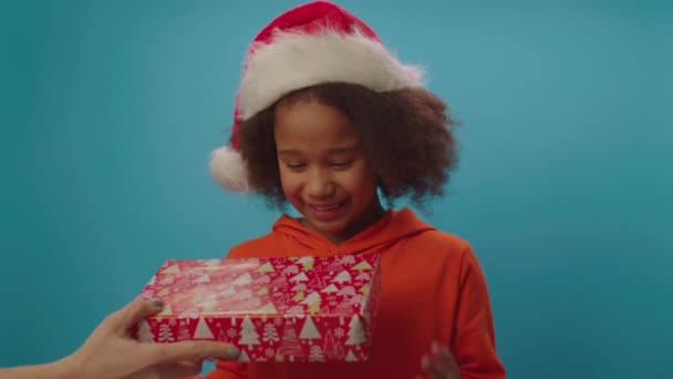 Menina afro-americana bonito em chapéu Santas recebe presente de Natal. Menina da escola feliz recebe presente de Natal em pé no fundo azul. — Vídeo de Stock