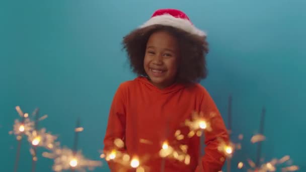 Menina afro-americana feliz em Santas chapéu dançando de pé sobre fundo azul com brilhos de Natal iluminação na frente. Miúdo saído ansioso pelo feriado de Natal. — Vídeo de Stock