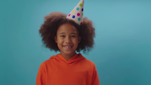 Sonriente chica afroamericana en sombrero de fiesta de cumpleaños hablando con la cámara de pie sobre fondo azul. Feliz niño felicitando con cumpleaños mirando a la cámara. Cantando feliz cumpleaños canción. — Vídeo de stock